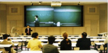 大学：正面の黒板と講師は、遠隔校舎からライブ講義を大型映像に映したもの （黄色い上着、筆者）。